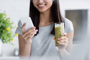 Elinde bir bardak smoothie ile ön planda akıllı telefondan mesaj atan esmer kadının kısmi görüntüsü