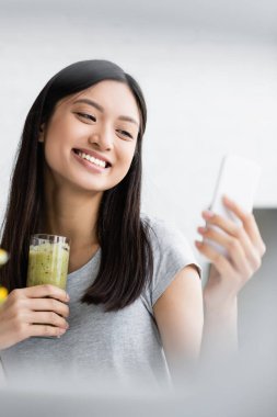 Neşeli Asyalı kadın bir bardak taze smoothie ile cep telefonundan selfie çekiyor.