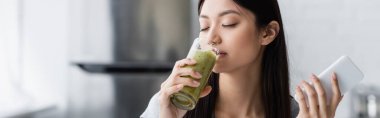 Akıllı telefonlu mutlu bir Asyalı kadın. Lezzetli bir smoothie içiyor, gözleri kapalı, pankart.