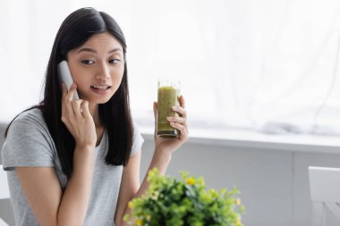 Genç Asyalı bir kadın cep telefonundan arıyor. Elinde bulanık bitkinin yanında bir bardak smoothie tutarken.