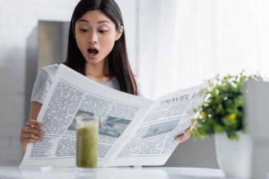 Şaşırmış Asyalı kadın seyahat gazetesi okuyor. Smoothie ile bulanık camların yanında.