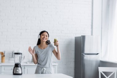Kulaklıklı mutlu Asyalı kadın bir bardak smoothie tutuyor ve el sallıyor.