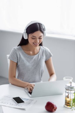 Kulaklıkla gülümseyen Asyalı kadın laptopta çaydanlığın yanında yazı yazıyor ve masanın üstünde akıllı telefon var.