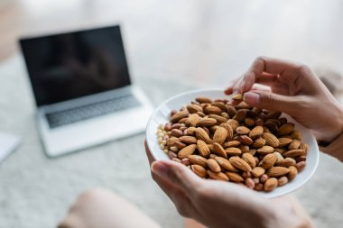 partial view of woman with bowl of almonds near blurred laptop with blank screen clipart