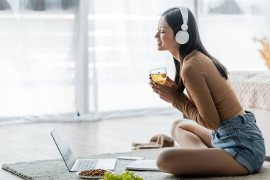 Kulaklıklı mutlu Asyalı kadın dizüstü bilgisayarının yanında çay ve yerde aperatif tutuyor.
