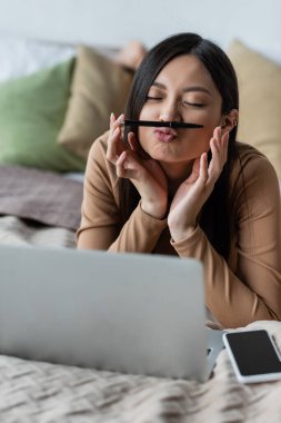 Burnu ve dudakları arasında dolma kalemle bulanık dizüstü bilgisayar ve akıllı telefonun yanında uzanan Asyalı bir kadın.
