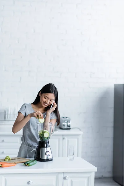 快乐的亚洲女人一边用手机聊天一边用搅拌机准备蔬菜 — 图库照片