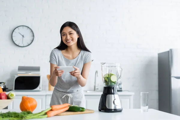 快乐的亚洲女人在厨房靠近电动摇椅的地方用手机拍照新鲜蔬菜 — 图库照片