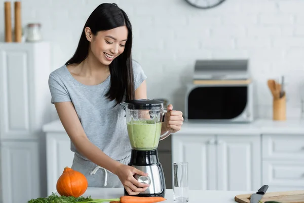 テーブルの上で熟したカボチャの近くのブレンダーで新鮮な野菜スムージーを準備笑顔アジアの女性 — ストック写真