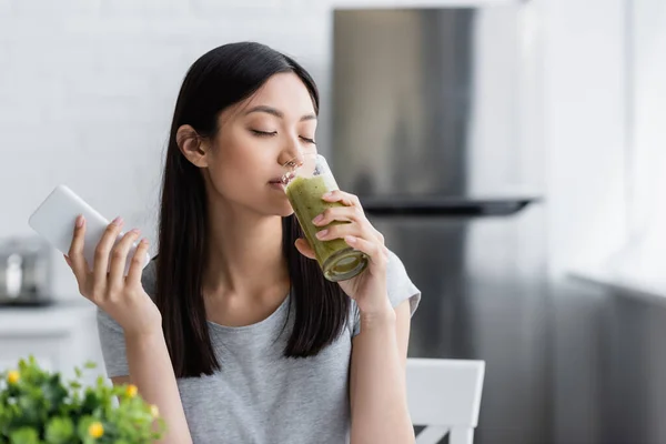 Satisfeito Asiático Mulher Com Celular Beber Delicioso Smoothie Cozinha — Fotografia de Stock