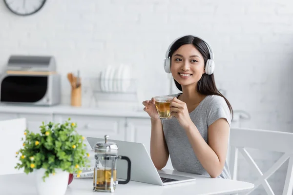 快乐的亚洲女人 头戴耳机 在笔记本电脑旁边端着一杯茶 在厨房里挂着模糊的植物 — 图库照片