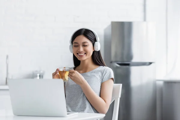 台所でノートパソコンを見ながらお茶を飲んでいるアジア系の陽気な女性 — ストック写真
