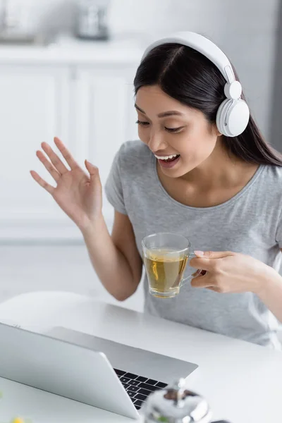 兴奋的亚洲女人 戴着耳机 一边举杯茶 一边在视频通话中挥手 — 图库照片