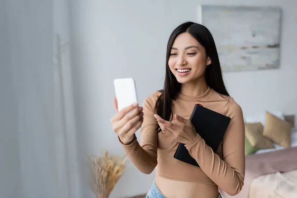 Glimlachende Aziatische Vrouw Met Kopieerboek Wijzend Met Vinger Tijdens Videogesprek — Stockfoto