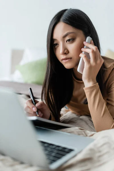 Yatakta Cep Telefonuyla Konuşurken Bulanık Dizüstü Bilgisayara Bakan Asyalı Kadın — Stok fotoğraf