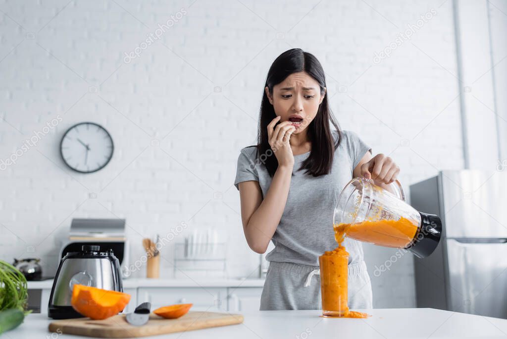 worried asian woman pouring smoothie into overflowing glass near cut pumpkin on chopping board