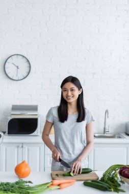 Neşeli Asyalı kadın taze sebzelerden kahvaltı hazırlarken taze salatalık kesiyor.