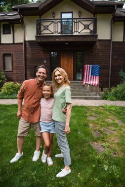 Tatil evindeki Amerikan bayrağının yanındaki kameraya gülümseyen aile. 