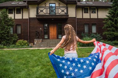 Back view of girl holding american flag while running on lawn near house  clipart