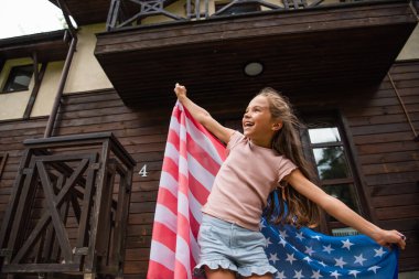 Amerikan bayrağını evin yanında tutan gülümseyen kızın alçak açılı görüntüsü 
