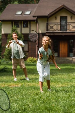 Gülümseyen kız çimenlikte akıllı telefonlu bulanık bir babanın yanında badminton oynuyor. 