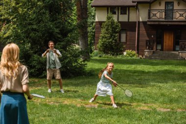 Anne ve çocuk açık havada babalarının yanında badminton oynuyorlar. 