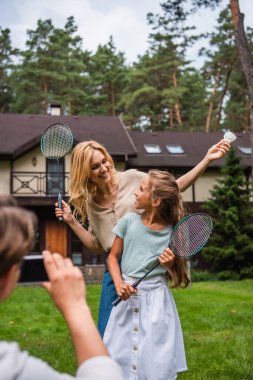 Elinde badminton raketi ve siki tutan gülümseyen kadın akıllı telefonlu bir eş ve kızının yanında. 