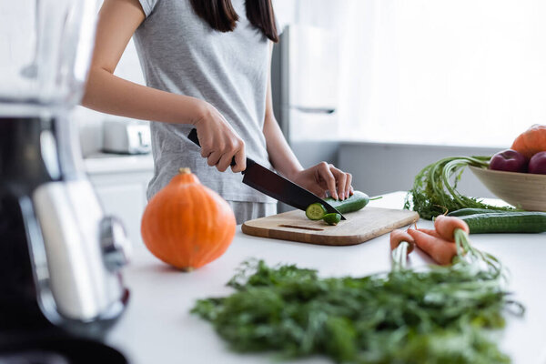 partial view of woman cutting cucumber near pumpkin and carrots on blurred foreground