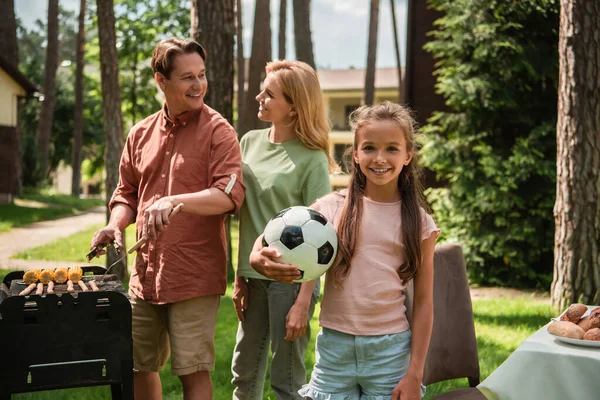 Sonriente Niño Con Fútbol Mirando Cámara Cerca Los Padres Cocinando — Foto de Stock