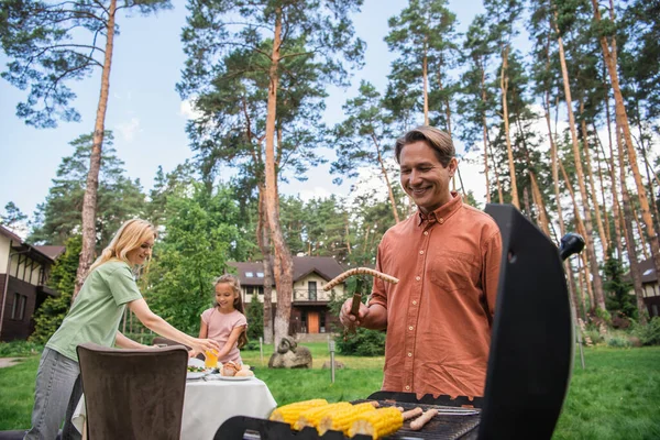 Smiling Man Holding Sausage Grill Blurred Family Outdoors — Stock fotografie