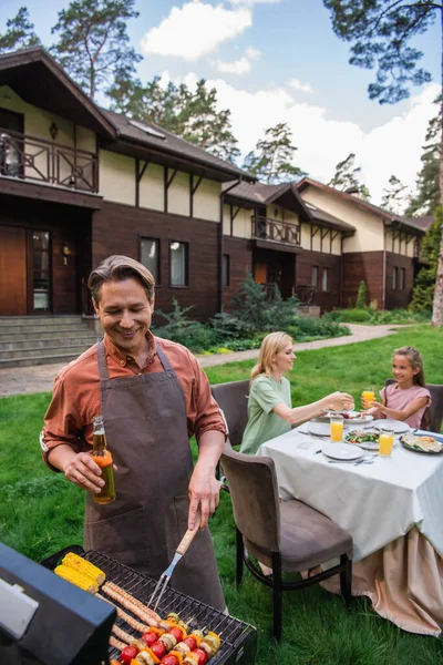 Smiling man with beer cooking food on grill near blurred family and vacation house