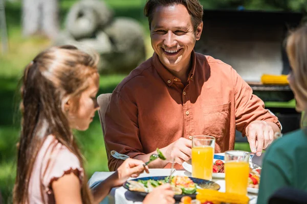 一个快乐的男人在外面的桌子上看着食物和橙汁旁边模糊的女儿 — 图库照片