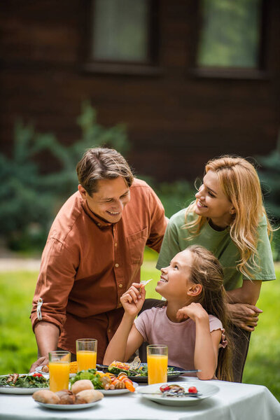 Smiling parents looking at kid near tasty food on table outdoors 