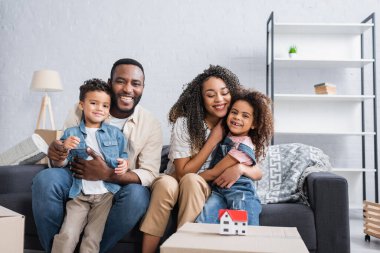 Afro-Amerikan ailesi karton kutunun üzerinde ev modelinin yanındaki koltukta oturuyor.