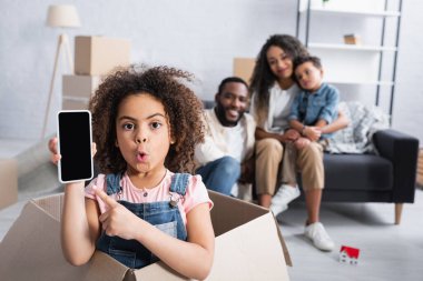 Karton kutudaki afro-amerikan kız akıllı telefonu işaret ediyor boş ekranla, bulanık ailenin yanında.