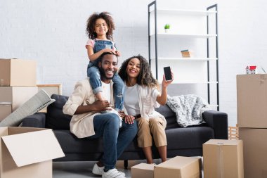 Neşeli Afrikalı Amerikalı kadın yeni dairede ailesinin yanında cep telefonunu boş ekranla gösteriyor. 
