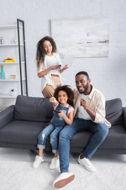 Afro-Amerikan baba ve kızı parmaklarıyla işaret ederken annesinin yanında dizüstü bilgisayarla televizyon izliyor.