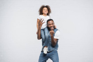 overjoyed african american man with happy daughter on shoulder showing yeah gesture on grey clipart