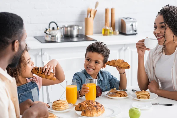 アフリカ系アメリカ人の少年は家族と朝食中にクロワッサンを持ちながら笑顔で — ストック写真