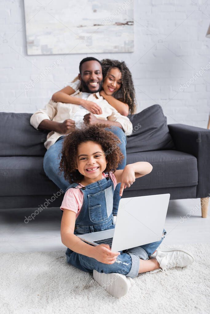 happy african american girl pointing at laptop while sitting on floor near blurred parents