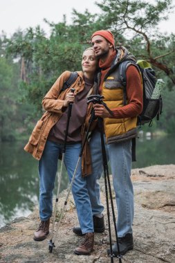 full length of happy woman holding hiking sticks and leaning on boyfriend with backpack 