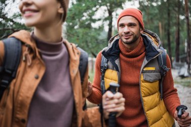 young man holding hiking stick and looking at happy girlfriend on blurred foreground