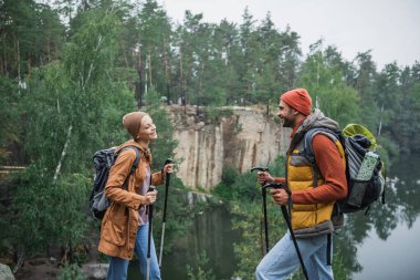happy couple with backpacks holding hiking sticks and looking at each other near lake clipart