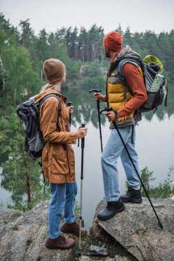 back view of couple with backpacks holding hiking sticks and looking at lake