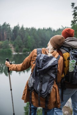 back view of woman with backpack holding hiking stick and leaning on man near at lake