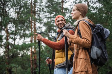 happy man pointing away with finger while looking at girlfriend with hiking sticks in forest 