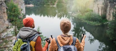 back view of couple with backpacks near lake in forest, banner