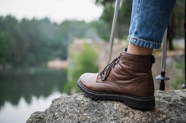 cropped view of woman in boot with hiking sticks standing on rock