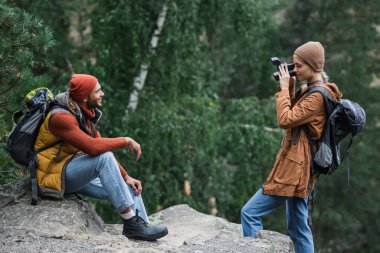 woman with backpack taking photo of happy boyfriend on vintage camera in forest 