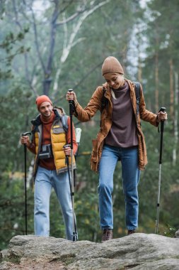 blurred man with vintage camera looking at smiling woman holding hiking sticks while trekking in forest